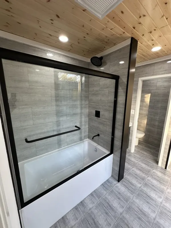Glass-enclosed tub and shower with grey tile under a pine ceiling