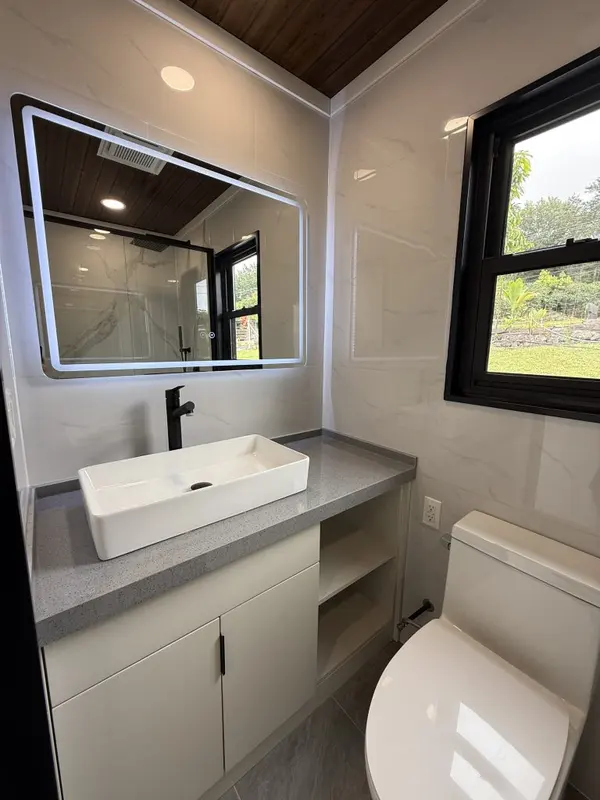 Bathroom with a white vessel sink, a lit mirror and a window, under a dark wood ceiling