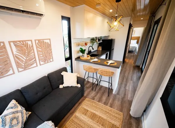 Living area with a charcoal sofa, palm-leaf wall art and a kitchen bar with rattan stools